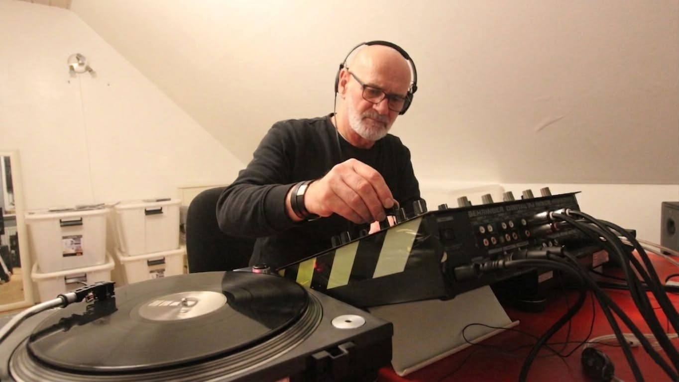 A bald, bearded man with glasses and headphones adjusting a knob on a DJ mixer next to a turntable
